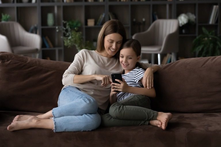 Happy kid and mom relaxing on sofa at home, using app on mobile phone, making video call, playing online game, shopping on internet together, browsing social media. Mother and girl looking at screen