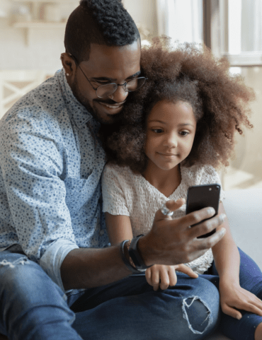 Loving young biracial dad sit on couch with cute little daughter watch funny video on smartphone together, happy caring african American father and small girl child use cellphone gadget at home