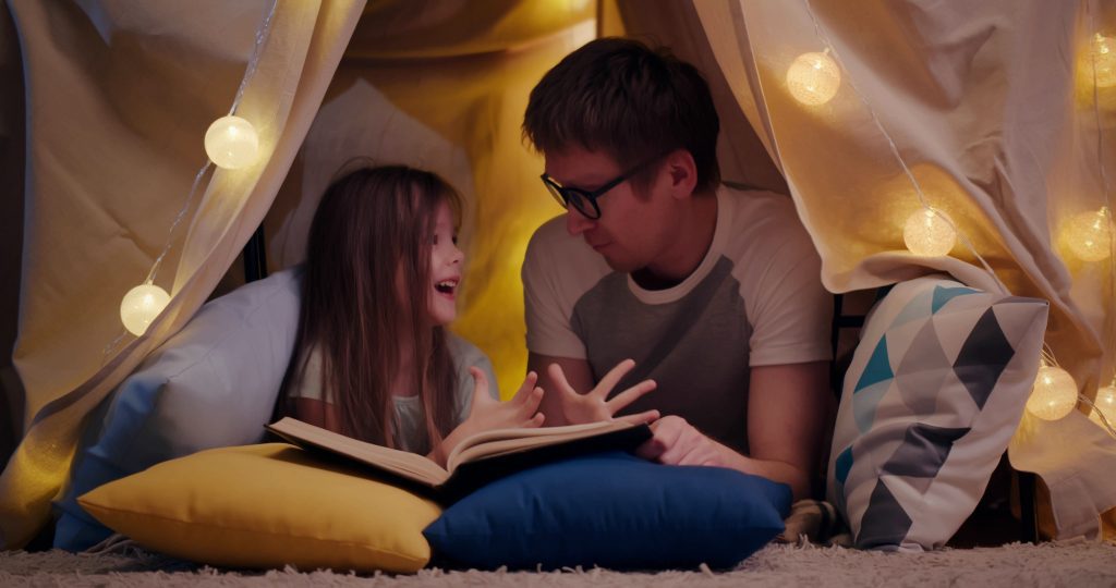 Happy father and daughter lying on floor in teepee and reading book together at home